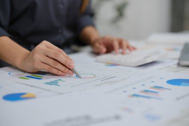Bir kadın masanın üstünde kalemle bir grafik üzerinde çalışıyor. Sayılara odaklanmış ve verileri analiz ediyor gibi görünüyor.