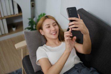 Bir kadın kanepede oturuyor ve telefonuyla kendi fotoğrafını çekiyor. Gülümsüyor ve anın tadını çıkarıyor.