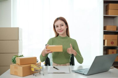 Bir kadın dizüstü bilgisayarı ve karton kutuları olan bir masada oturuyor. Elinde bir karton kutu tutuyor ve ona başparmak işareti yapıyor.