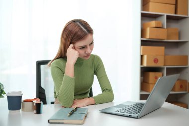 Masasında dizüstü bilgisayarı ve not defteri olan bir kadın oturuyor. Kaşlarını çatıyor ve bilgisayar ekranına bakıyor.