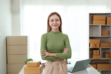 Bir kadın elinde dizüstü bilgisayar ve bir yığın karton kutuyla bir masanın önünde duruyor. Yeşil bir gömlek giyiyor ve kollarını kavuşturuyor. Sahne profesyonel ya da iş ortamı öneriyor.