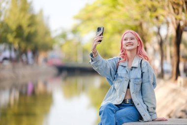 Pembe saçlı bir kadın cep telefonuyla selfie çekiyor. Bir su kütlesinin yanındaki çıkıntıda oturuyor.