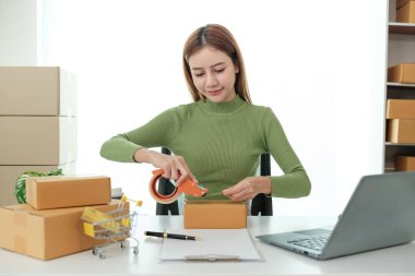 Bir kadın dizüstü bilgisayarı ve karton kutuları olan bir masada oturuyor. Kutuları mühürlemek için bant kullanıyor.