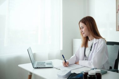 Beyaz laboratuvar önlüklü bir kadın dizüstü bilgisayarı ve kalemi olan bir masada oturuyor. Bir kağıt parçasına bir şeyler yazıyor.