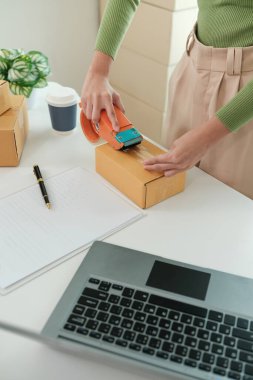 Bir kadın karton bir kutuyu mühürlemek için bant kesicisi kullanıyor. Kutu bir dizüstü bilgisayarın yanındaki masada.
