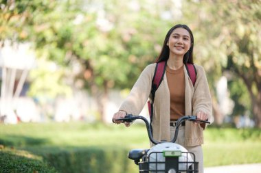 Önünde sepeti olan bir kadın bisiklete biniyor. Gülümsüyor ve sırt çantası takıyor.