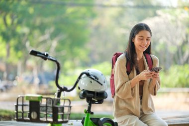 Yanında bir kask ve bisikletle bankta oturan bir kadın var. Telefonuna bakıyor.