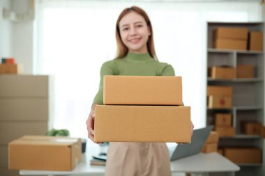 Bir kadın iki kutu tutuyor ve gülümsüyor. Kutular kahverengi ve karton gibi görünüyor. Kadın üstünde dizüstü bilgisayarla bir masanın önünde duruyor.