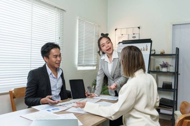 Üç kişi bir iş ortamında bir masada oturuyor. Adamlardan biri dizüstü bilgisayar tutuyor, diğer ikisi de ona bakıyor. Sağdaki kadın beyaz bir bluz giyiyor.