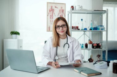 Beyaz laboratuvar önlüklü bir kadın elinde dizüstü bilgisayarla bir masada oturuyor. Gülümsüyor ve bir doktor.