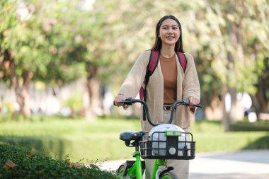 Önünde sepeti olan bir kadın bisiklete biniyor. Gülümsüyor ve sırt çantası takıyor.