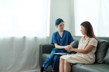 Mavi önlüklü bir kadın bir kanepede başka bir kadınla oturuyor. Mavi önlüklü kadın diğer kadınların elini tutuyor.