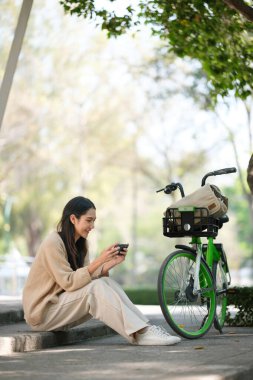 Bir kadın, yanında yeşil bir bisikletle merdivende oturuyor. Telefonuna bakıyor.
