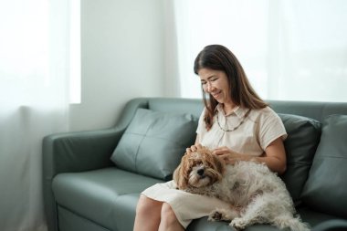 Bir kadın beyaz bir köpekle kanepede oturuyor. Gülümsüyor ve köpeği okşuyor.