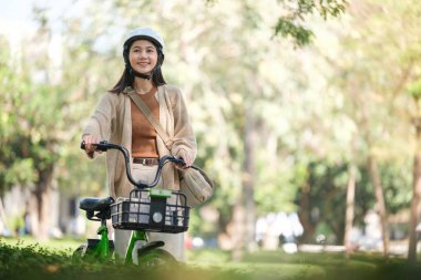 Önünde sepeti olan bir kadın bisiklete biniyor. O bir kask ve kahverengi ceket giyiyor.