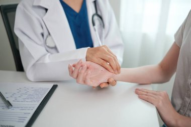 Bir doktor bir hastanın elini muayene ediyor. Hasta beyaz bir gömlek giyiyor.