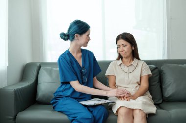 Hemşirenin biri bir hastayla kanepede oturuyor. Hemşire mavi bir gömlek giyiyor ve hasta beyaz bir gömlek giyiyor. Hemşire hastaların elini tutuyor. Rahatlık ve destek sağlıyor.