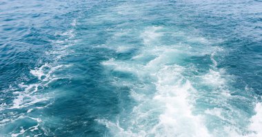 Teknenin arkasında ve deniz yüzeyinin dokusunda beyaz köpüklü dalgalar oluşuyor..
