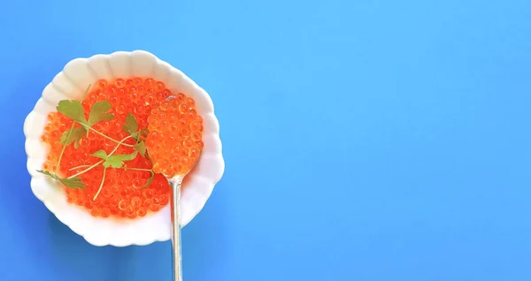 Red caviar and parsley in spoon on white plate. Caviar in bowl on blue background. Close-up salmon caviar. Caviar texture. Seafood background