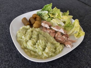 Beyaz tabakta servis edilen ev yapımı ızgara tavuk, kabak güveci, yeşil salata ve salatalık turşusu.