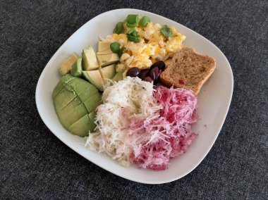 Avokado, omlet, zeytin, kohlrabi, kırmızımsı ve beyaz bir tabakta servis edilen bir dilim ekmek ile ev yapımı kahvaltı.