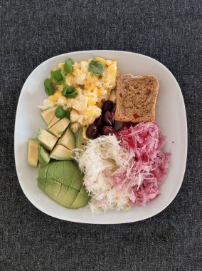 Avokado, omlet, zeytin, kohlrabi, kırmızımsı ve beyaz bir tabakta servis edilen bir dilim ekmek ile ev yapımı kahvaltı.