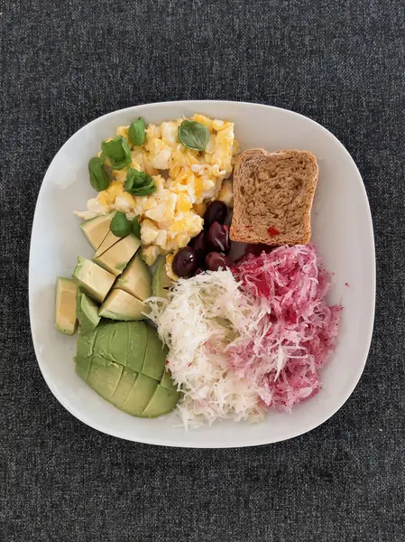 Avokado, omlet, zeytin, kohlrabi, kırmızımsı ve beyaz bir tabakta servis edilen bir dilim ekmek ile ev yapımı kahvaltı.