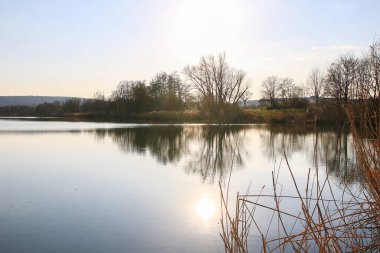 Sakin bir bahar sahnesi, çıplak ağaçlarla sakin bir göl ve suda güzel bir şekilde yansıyan berrak bir gökyüzü..