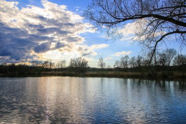 Sakin bir bahar sahnesi, çıplak ağaçlarla sakin bir göl ve suda güzel bir şekilde yansıyan berrak bir gökyüzü..