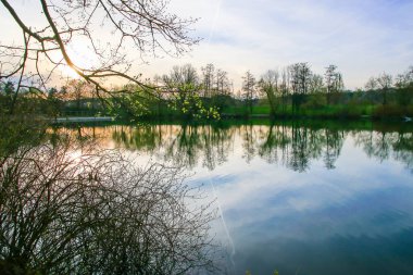 Sakin bir bahar sahnesi, çıplak ağaçlarla sakin bir göl ve suda güzel bir şekilde yansıyan berrak bir gökyüzü..