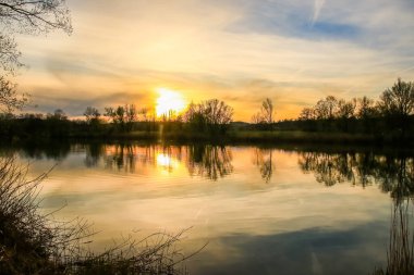 Sakin bir bahar sahnesi, çıplak ağaçlarla sakin bir göl ve suda güzel bir şekilde yansıyan berrak bir gökyüzü..