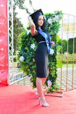 TIMISOARA, ROMANIA - 12 Temmuz 2022: Uyumlu siyah bir elbise giyen kendinden emin genç bir mezun üniversite mezuniyetinde akademik başarısını kutlarken gururla gülümsüyor