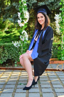 TIMISOARA, ROMANIA - 12 Temmuz 2022: Yüksek topuklu giyen genç çekici bayan öğrenci mezuniyet kutlaması sırasında çiçek salıncağında zarif bir şekilde gülümsüyor