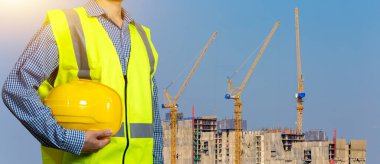 Güvenlik giysisi konsepti. Sarı kaskı ya da kaskı tutan erkek eli. Yansıtıcı sarı yelekli inşaat işçisi tamamlanmamış binanın önünde duruyor.