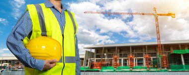 Güvenlik giysisi konsepti. Sarı kaskı ya da kaskı tutan erkek eli. Yansıtıcı sarı yelekli inşaat işçisi tamamlanmamış binanın önünde duruyor.