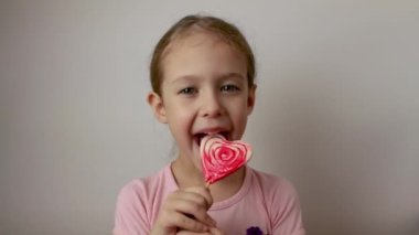Sevimli küçük bir kız kalp şeklindeki lolipopu emiyor. Sevgililer Günü. İzole edilmiş beyaz arkaplan.
