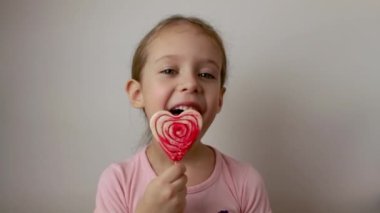 Happy girl licks a delicious red heart shaped lollipop. Valentines Day. Isolated white background.