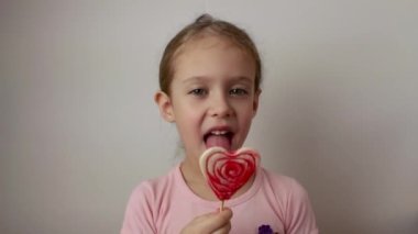 A cute little girl licks a big striped heart-shaped lollipop. Isolated white background. Valentines Day.