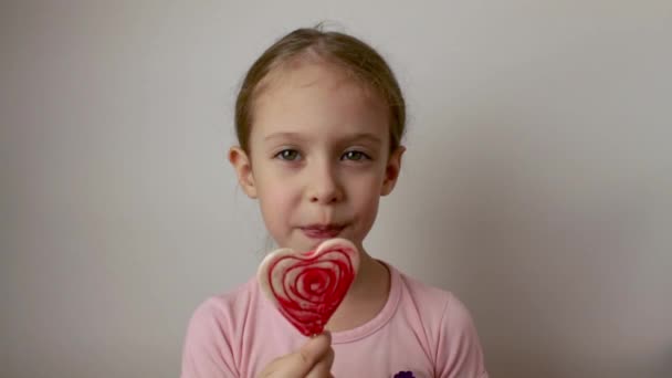 幸せな女の子はおいしいハート型のロリポップを舐めます バレンタインデー 隔離された白い背景 — ストック動画