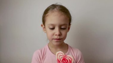 A cute little girl licks a heart-shaped lollipop. Valentines Day. Delicious dessert. Isolated white background.