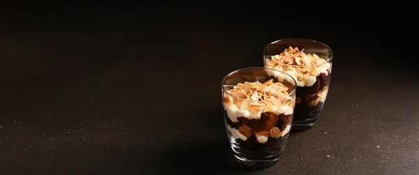 Chocolate cake in a glass with vanilla cream and almonds. A portion of dessert in a glass on a dark background.