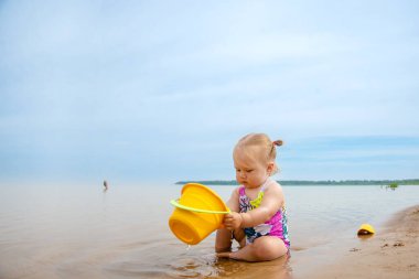 Mayo giymiş küçük bir kız yazın göl kenarında kovayla ve kumla oynar.