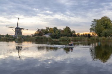 Tipik bir Hollanda manzarası - bir kürekçi sabah erken saatlerde eski bir yel değirmeni olan Amstel nehri boyunca yüzer.
