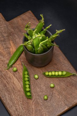 Seramik bardakta yeşil bezelye kabukları ve karanlık masadaki ahşap tahtaya serpiştirilmiş.