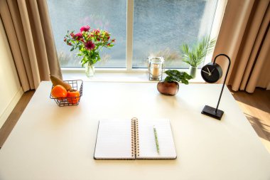 Beyaz bir masanın üzerinde temiz bir defter ve bir kalem var. Masa rezervuara bakan büyük bir pencerenin yanında duruyor. Pencere dışında ilham verici bir görünüm planlama ya da günlük girişi için atmosfer yaratır