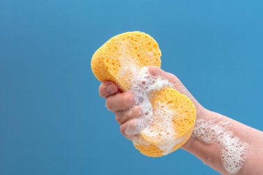 El, mavi arka planda sabunlu köpüklü sarı bir süngeri sıkıca tutar..