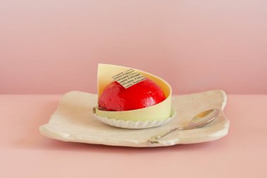 Dekorlu kırmızı bir pastanenin yakın plan fotoğrafı kare seramik bir tabağa sergilenmiş ve arka planda pembe bir kaşık var..  