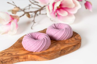 Beyaz bir masanın üzerindeki ahşap dokuma tahtasının üzerinde iki tane köpük pembesi. Arkasında bir manolya filizi yatıyor.