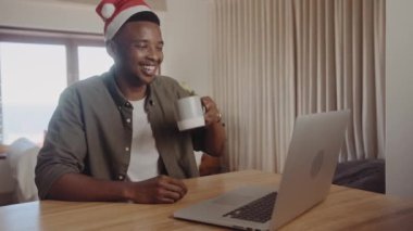 Genç Afrikalı siyahi erkek Noel şapkası takıyor ve ailesine ve arkadaşlarına bir internet video görüşmesinde kahve içerken neşeli bir sezon diliyorlar. Mutfak tezgahında dizüstü bilgisayar kullanarak oturmak.