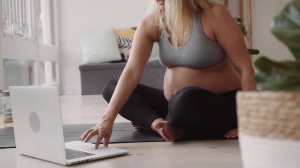 怀孕的白种人年轻女性 在客厅里坐在瑜伽垫上 在笔记本电脑上进行在线伸展教学 — 图库视频影像